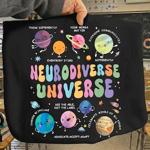 Könnte beinhalten: Schwarze Tragetasche mit einem farbenfrohen Design, das Planeten mit Gesichtern und den Schriftzug "Neurodiverse Universum" zeigt. Das Design enthält Sätze wie "Denke anders" und "Feiere alle Geister".
