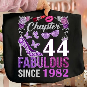 Puede incluir: Camiseta negra con la frase "Chapter 44 Fabulous Since 1982". El diseño incluye un zapato de tacón, mariposas, lápiz labial, gafas y detalles florales en morado y plateado.