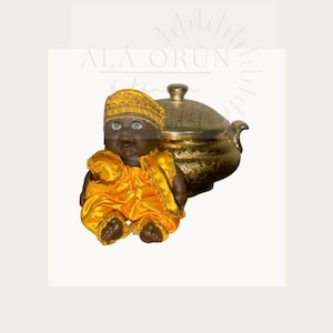 May include: A small black doll wearing a yellow satin outfit and a gold crown. The doll is sitting in front of a gold serving dish.