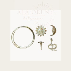 Op de afbeelding: Een set zilveren bedels met een zon, maan, slang, hand en een cirkel. De bedels zijn op een witte achtergrond met de tekst "ALÁ ORÚN botánica".