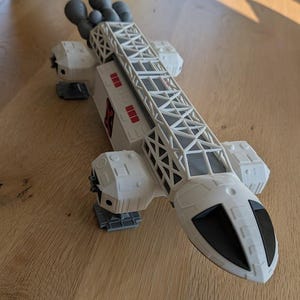 Pode incluir: Modelo de nave espacial branco e cinza com um design detalhado. O modelo apresenta uma longa estrutura em treliça no centro, com motores cinza e detalhes vermelhos. A nave espacial tem aproximadamente 46 cm de comprimento.