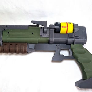Könnte beinhalten: Ein Sci-Fi-Requisiten-Blaster mit dunkelgrauem Rahmen, olivgrünem Lauf und Griff sowie einem braunen, gerippten Vorderschaft. Ein gelbes zylindrisches Bauteil mit roten Pfeilen sowie Plus- und Minuszeichen ist oben auf dem Blaster montiert.