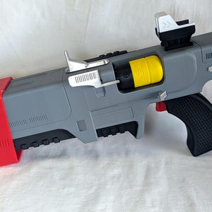 Puede incluir: Un bláster de juguete gris con una culata trasera de color rojo brillante, una empuñadura de pistola negra texturizada y un componente cilíndrico amarillo. El dispositivo incluye una palanca superior metálica plateada, una mira negra y blanca y un dial plateado circular.