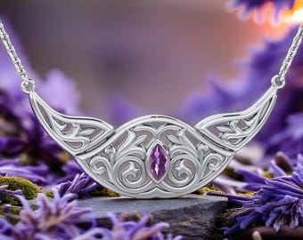 May include: A silver necklace with an intricate, ornate design featuring a purple gemstone in the center. The necklace is displayed on a background of purple flowers.