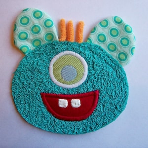 May include: A teal monster-shaped applique with a single eye, a red mouth, and two white teeth. The monster has two ears with a green polka dot pattern and orange antennae. The applique is made of a soft, textured material.