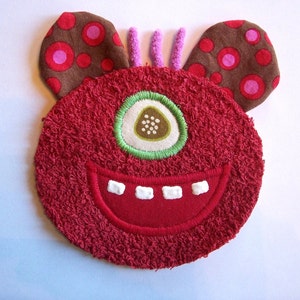 May include: A plush, red monster face with one large eye, a wide smile, and two brown ears with pink polka dots. The monster has three purple antennae. This whimsical design is suitable for children's decor or accessories.