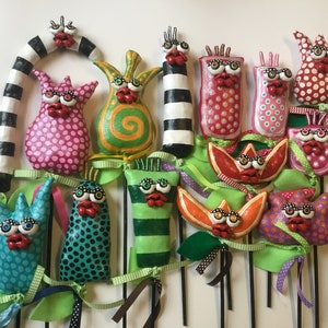 May include: A collection of whimsical, handmade, colorful, and patterned figures with googly eyes and red lips. The figures are attached to sticks and have green, pink, and purple ribbons.
