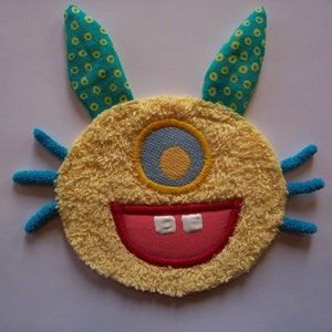 May include: A plush, yellow monster face with a single blue eye, a wide red mouth, and two white teeth. It has blue antennae and green and yellow patterned ears. The design is likely for a child's toy or decorative item.