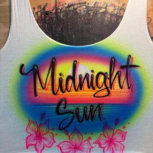 May include: A white tank top featuring an airbrushed design of a sunset with a rainbow-colored aura. The text Midnight Sun is written in black cursive script across the center. Three pink hibiscus flowers are painted along the bottom edge.