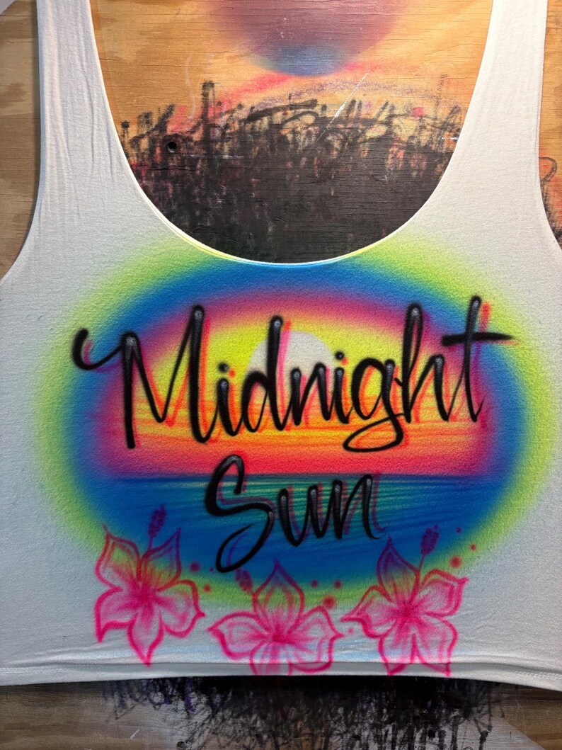 May include: A white tank top featuring an airbrushed design of a sunset with a rainbow-colored aura. The text Midnight Sun is written in black cursive script across the center. Three pink hibiscus flowers are painted along the bottom edge.