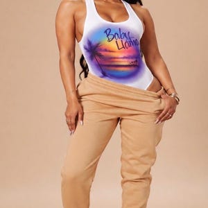 May include: A white racerback bodysuit featuring a colorful airbrushed sunset graphic with palm trees and the text Baby Llama, paired with tan sweatpants and white sneakers. The bodysuit has a scoop neckline and a fitted silhouette.