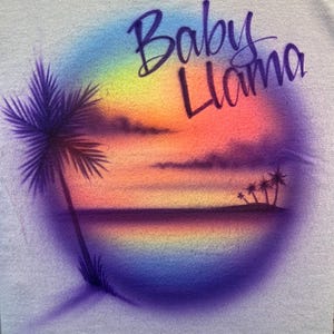 May include: A light-colored fabric featuring an airbrushed circular design of a tropical sunset with a palm tree on the left and smaller palm trees on the right. The text Baby Llama is written in a dark purple cursive script across the top.