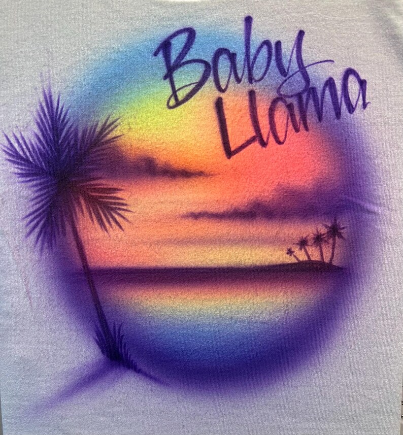 May include: A light-colored fabric featuring an airbrushed circular design of a tropical sunset with a palm tree on the left and smaller palm trees on the right. The text Baby Llama is written in a dark purple cursive script across the top.