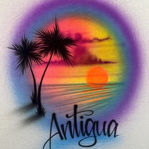 May include: A white t-shirt featuring an airbrushed sunset scene with two palm trees on the left, a bright orange sun over the water, and a vibrant purple, pink, and yellow gradient background. The word Antigua is written in black script below.