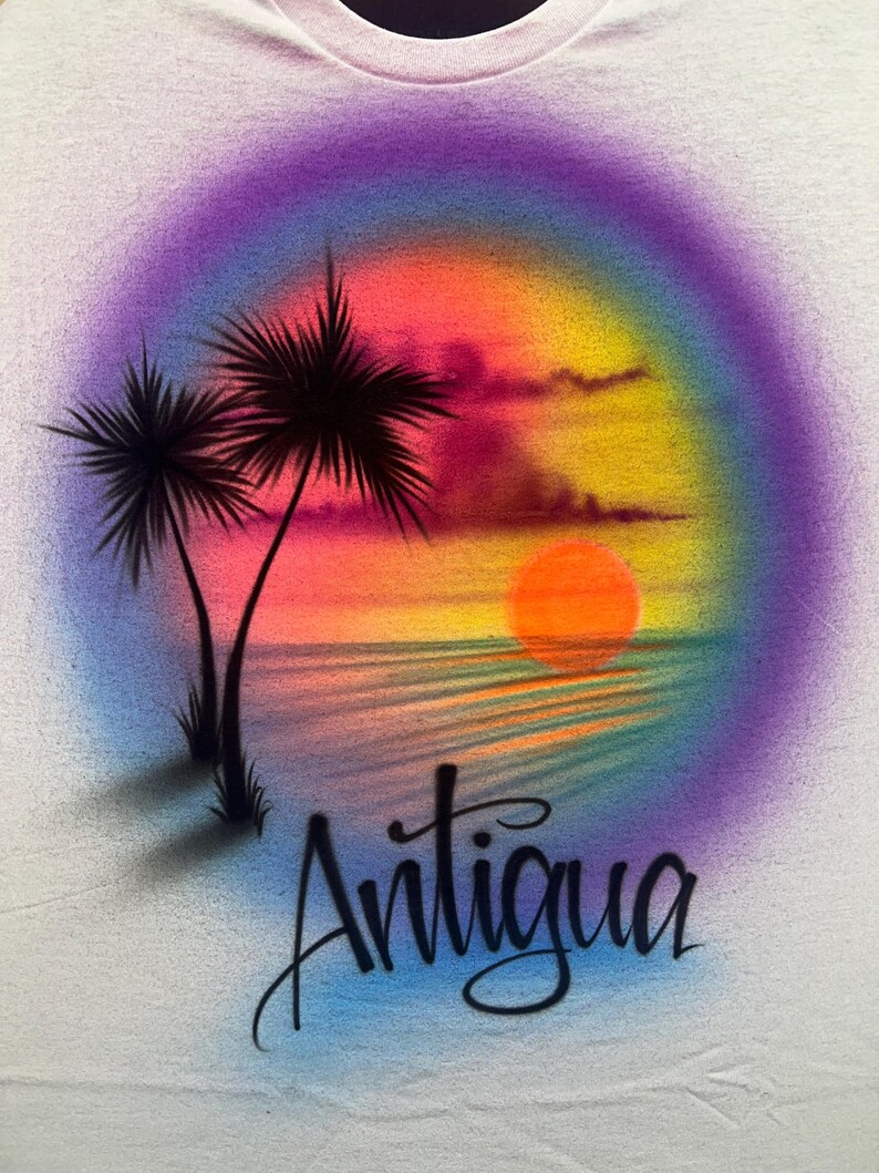 May include: A white t-shirt featuring an airbrushed sunset scene with two palm trees on the left, a bright orange sun over the water, and a vibrant purple, pink, and yellow gradient background. The word Antigua is written in black script below.