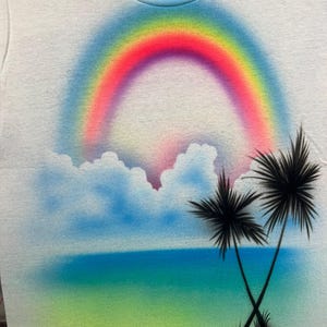 May include: A white t-shirt featuring an airbrushed graphic of a vibrant rainbow arching over fluffy blue clouds. Below the clouds, a gradient of blue and green represents the ocean and shore, with two black palm trees silhouetted in the foreground.