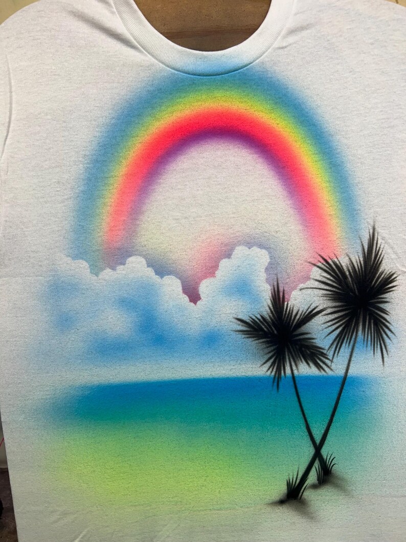 May include: A white t-shirt featuring an airbrushed graphic of a vibrant rainbow arching over fluffy blue clouds. Below the clouds, a gradient of blue and green represents the ocean and shore, with two black palm trees silhouetted in the foreground.