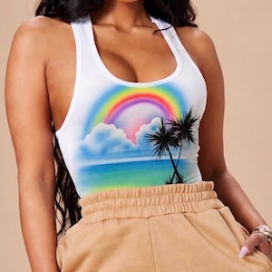 May include: A white racerback tank top featuring a colorful airbrushed graphic of a rainbow, fluffy clouds, a blue ocean horizon, and black palm tree silhouettes, paired with tan high-waisted sweatpants with an elasticized waistband.