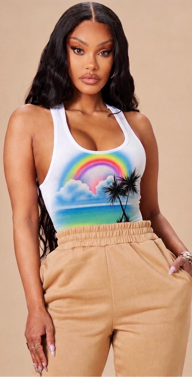 May include: A white racerback tank top featuring a colorful airbrushed graphic of a rainbow, fluffy clouds, a blue ocean horizon, and black palm tree silhouettes, paired with tan high-waisted sweatpants with an elasticized waistband.