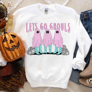 May include: White sweatshirt with the text "Lets Go Ghouls" in pink. The graphic features three ghosts in pink sheets, turquoise boots, and black eyes. The ghosts stand on a bed of silver glitter. A pumpkin and fall decor are in the background.