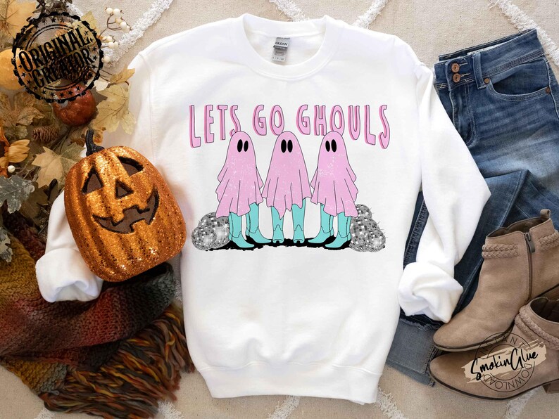 May include: White sweatshirt with the text "Lets Go Ghouls" in pink. The graphic features three ghosts in pink sheets, turquoise boots, and black eyes. The ghosts stand on a bed of silver glitter. A pumpkin and fall decor are in the background.