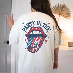 K&ouml;nnte beinhalten: Wei&szlig;es T-Shirt mit einem Grafikdruck einer roten, wei&szlig;en und blauen Zunge, die herausstreckt, mit den Worten "Party in the USA" auf der R&uuml;ckseite.