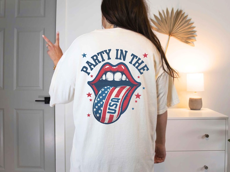 K&ouml;nnte beinhalten: Wei&szlig;es T-Shirt mit einem Grafikdruck einer roten, wei&szlig;en und blauen Zunge, die herausstreckt, mit den Worten "Party in the USA" auf der R&uuml;ckseite.