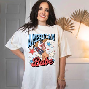Puede incluir: Una camiseta blanca con un estampado gr&aacute;fico de una mujer con botas de vaquero con estrellas rojas, blancas y azules y el texto "American Babe".