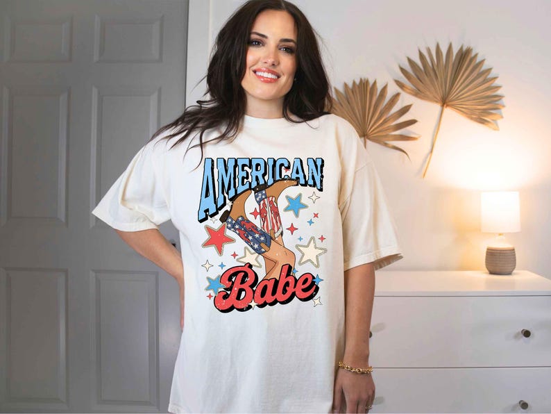 Puede incluir: Una camiseta blanca con un estampado gr&aacute;fico de una mujer con botas de vaquero con estrellas rojas, blancas y azules y el texto "American Babe".