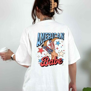 Puede incluir: Camiseta blanca con un estampado gr&aacute;fico de una mujer con botas de vaquero con estrellas y el texto "American Babe".
