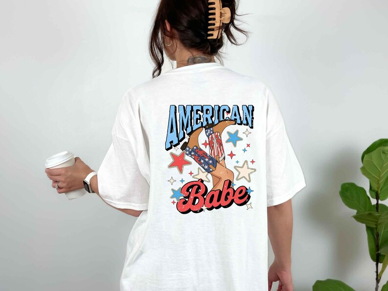 Puede incluir: Camiseta blanca con un estampado gr&aacute;fico de una mujer con botas de vaquero con estrellas y el texto "American Babe".