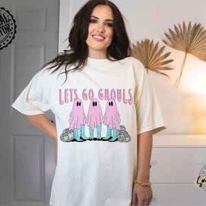 May include: Cream-colored t-shirt with the phrase "Lets Go Ghouls" in pink lettering. The shirt features a graphic of three pink ghosts with black eyes and blue boots. The design is centered on the front of the shirt.