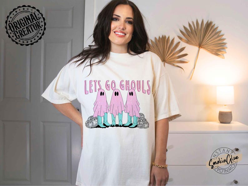 May include: Cream-colored t-shirt with the phrase "Lets Go Ghouls" in pink lettering. The shirt features a graphic of three pink ghosts with black eyes and blue boots. The design is centered on the front of the shirt.