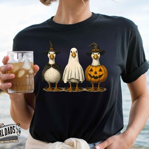 May include: Black t-shirt featuring three geese dressed for Halloween. One goose is in a witch costume, another is a ghost, and the third is a jack-o'-lantern. The shirt has a whimsical, cartoonish design, perfect for the spooky season.