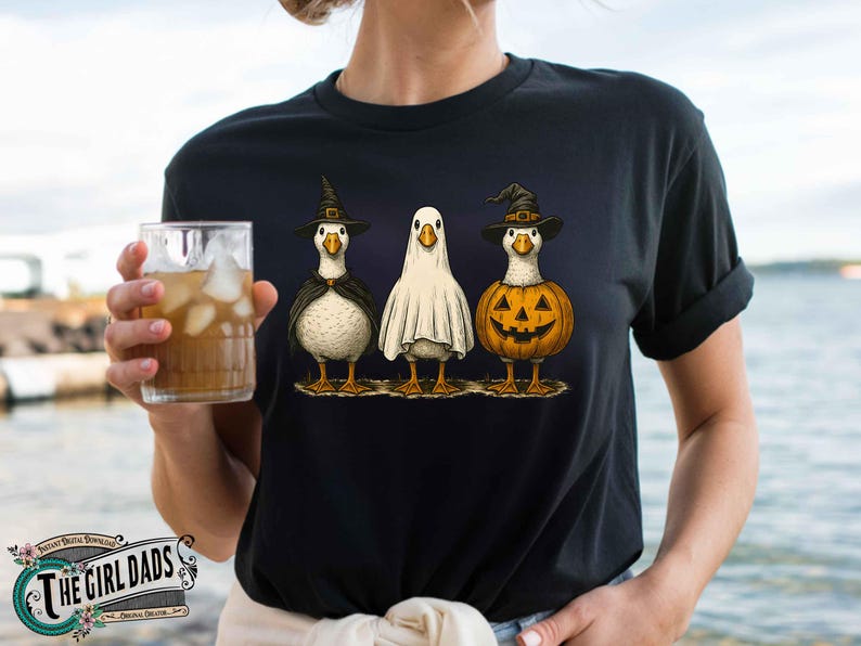 May include: Black t-shirt featuring three geese dressed for Halloween. One goose is in a witch costume, another is a ghost, and the third is a jack-o'-lantern. The shirt has a whimsical, cartoonish design, perfect for the spooky season.