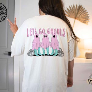 May include: Cream-colored t-shirt with the phrase "Lets Go Ghouls" in pink lettering. The shirt features a graphic of three pink ghost figures wearing turquoise boots. The design includes silver glitter accents.