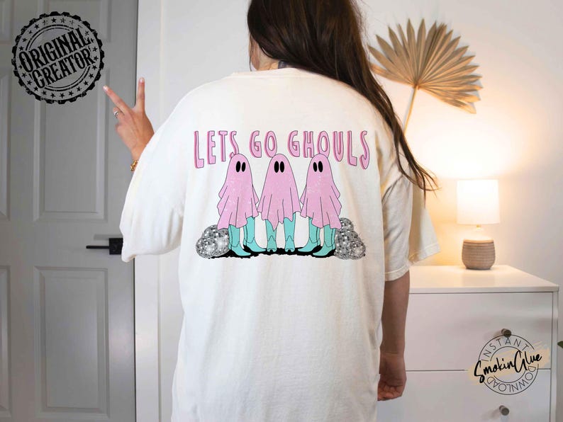 May include: Cream-colored t-shirt with the phrase "Lets Go Ghouls" in pink lettering. The shirt features a graphic of three pink ghost figures wearing turquoise boots. The design includes silver glitter accents.