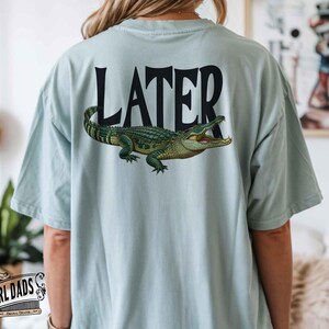 May include: Light blue t-shirt with the word "LATER" in black block letters and a detailed illustration of a green and yellow alligator. The shirt is a casual style.
