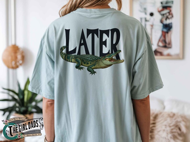 May include: Light blue t-shirt with the word "LATER" in black block letters and a detailed illustration of a green and yellow alligator. The shirt is a casual style.