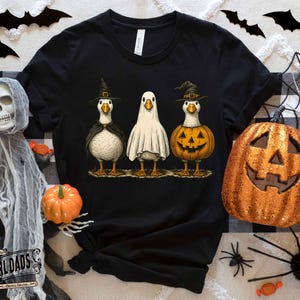 May include: Black t-shirt featuring three cartoon geese dressed for Halloween. One goose wears a witch hat and cape, another is a ghost, and the third is a jack-o'-lantern. Halloween-themed apparel.