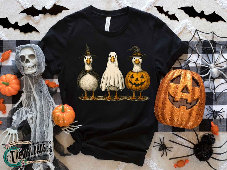 May include: Black t-shirt featuring three cartoon geese dressed for Halloween. One goose wears a witch hat and cape, another is a ghost, and the third is a jack-o'-lantern. Halloween-themed apparel.