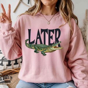 May include: A light pink sweatshirt with the word "LATER" in black block letters and a detailed illustration of a green and yellow alligator. The sweatshirt is a long-sleeved crew neck.