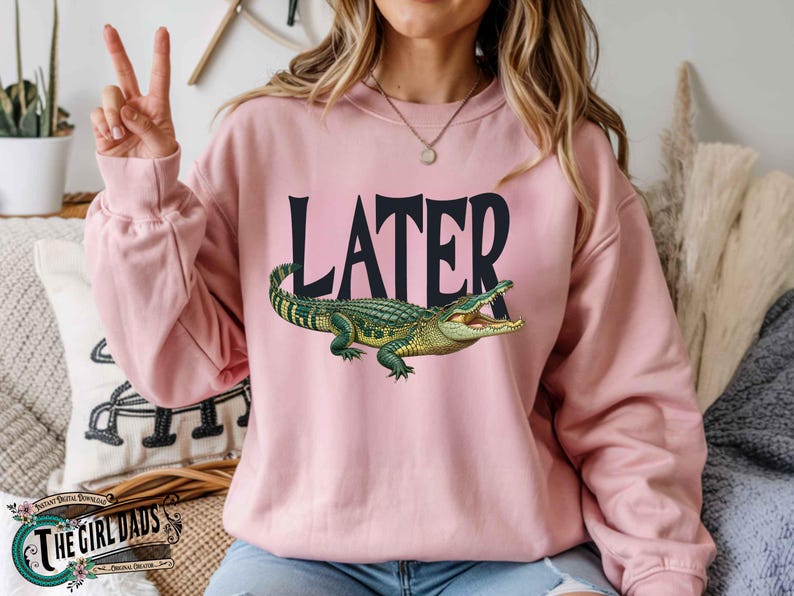 May include: A light pink sweatshirt with the word "LATER" in black block letters and a detailed illustration of a green and yellow alligator. The sweatshirt is a long-sleeved crew neck.