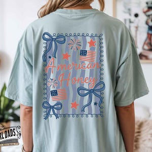 May include: Light blue t-shirt with a patriotic design. The back of the shirt features the text "American Honey" in a decorative frame with bows, stars, fireworks, and American flags. The shirt is a comfortable, casual style.