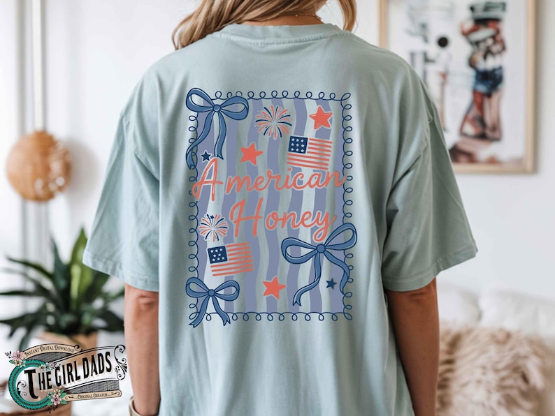 May include: Light blue t-shirt with a patriotic design. The back of the shirt features the text "American Honey" in a decorative frame with bows, stars, fireworks, and American flags. The shirt is a comfortable, casual style.