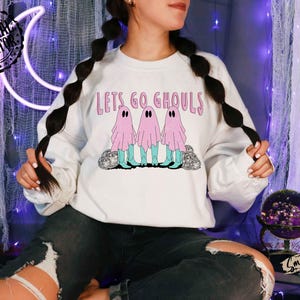 May include: White sweatshirt with the text "Lets Go Ghouls" in pink. The graphic features three pink ghost figures with teal boots. The sweatshirt is a Halloween-themed item.