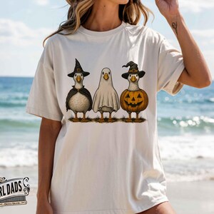 May include: Beige t-shirt featuring three cartoon geese dressed for Halloween. One goose wears a witch hat and cape, another is a ghost, and the third is a jack-o'-lantern. The Girl Dads logo is at the bottom.