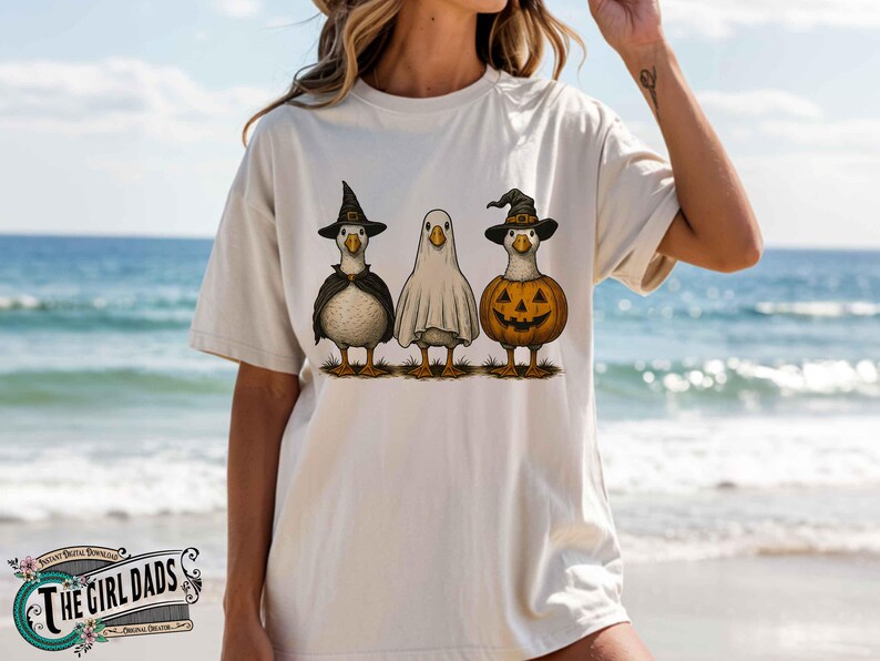 May include: Beige t-shirt featuring three cartoon geese dressed for Halloween. One goose wears a witch hat and cape, another is a ghost, and the third is a jack-o'-lantern. The Girl Dads logo is at the bottom.