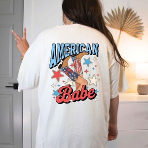 Puede incluir: Camiseta blanca con un gr&aacute;fico de una mujer con botas de vaquero y el texto "American Babe" en azul y rojo.