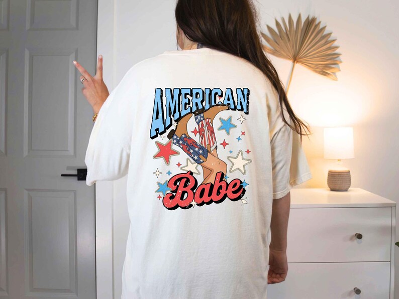 Puede incluir: Camiseta blanca con un gr&aacute;fico de una mujer con botas de vaquero y el texto "American Babe" en azul y rojo.